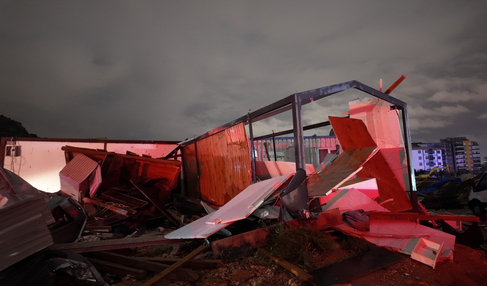 Gece çekilmiş fotoğrafta şantiye alanında yerle bir olmuş konteyner enkazı; metal iskelet, dağılmış panel ve tahta parçaları, arkada apartman blokları