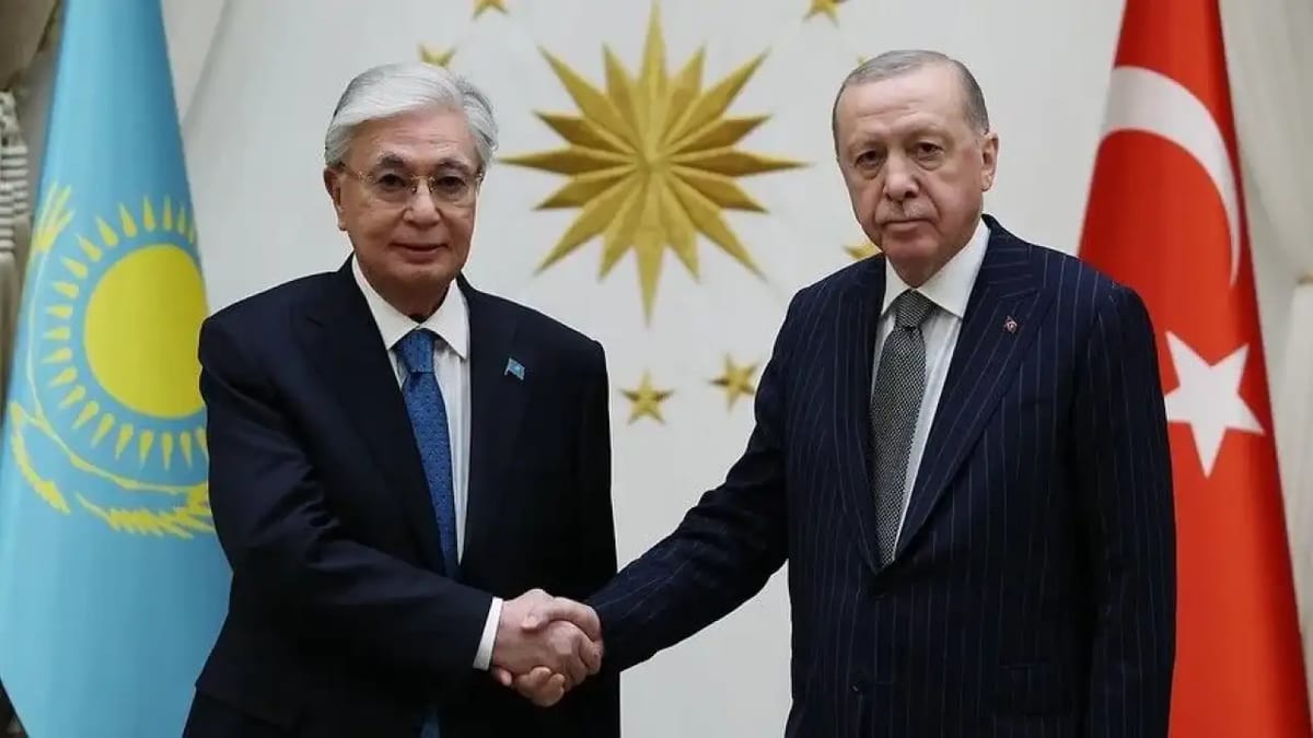 Cumhurbaşkanlığı armasının önünde, solda Kazakistan, sağda Türkiye bayrağıyla, koyu takım elbiseli iki erkek el sıkışıyor