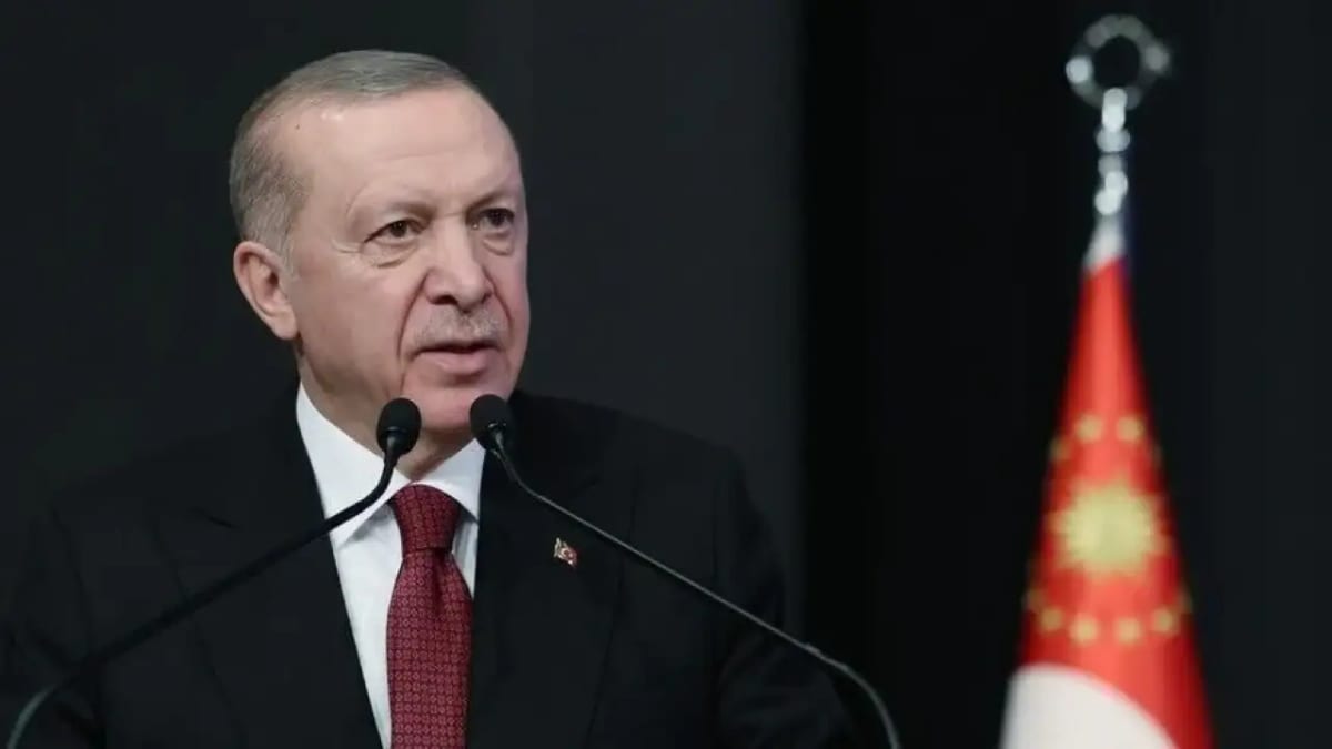 İki mikrofonun arkasında konuşan, koyu takım elbiseli ve kırmızı kravatlı bir erkek; sağda bulanık arka planda kırmızı bir Cumhurbaşkanlığı flaması
