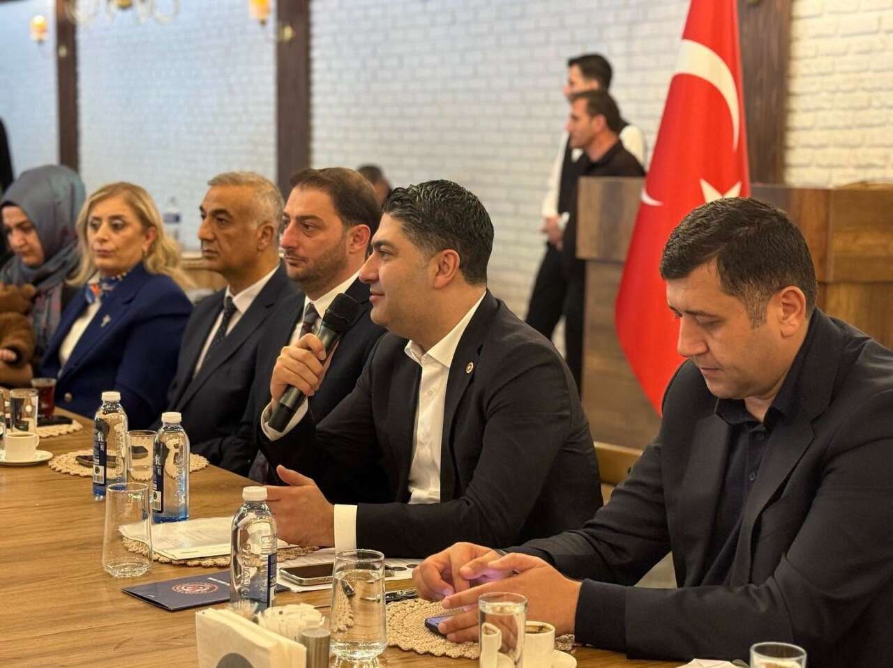 Uzun bir masanın başında oturan takım elbiseli erkekler ve kadınlar; ortadaki erkek mikrofonla konuşuyor, arkada Türk bayrağı var
