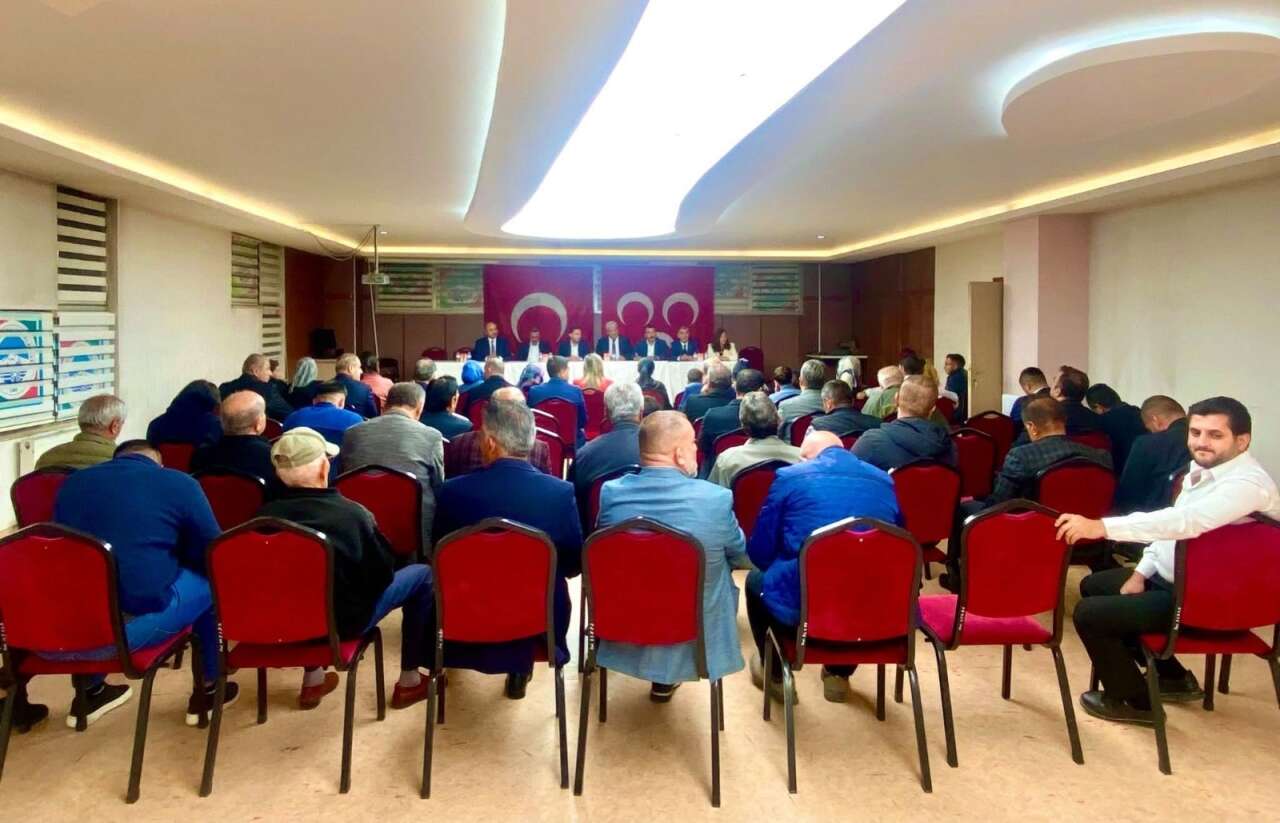 Kırmızı sandalyelerde oturan kalabalık bir topluluk, karşıda MHP bayraklarının önündeki uzun masada oturan yöneticiler
